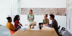 Four co-workers sitting in a bright and modern meeting room, with one woman standing, holding a tablet in her hands while talking to her colleagues.