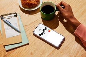 Phone displaying a PayPal Business Debit Mastercard in mobile wallet ready for contactless payment, next to a coffee mug, croissant, and a paid receipt on a clipboard.