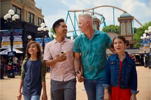 A joyful family of four walking through a theme park on a sunny day, smiling and laughing, with a roller coaster visible in the background.