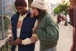 Two people standing on a sidewalk smiling at a smartphone.