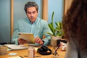 Hombre concentrado mirando su tablet, sentado en una mesa con otros objetos, como documentos, unos auriculares y una planta.