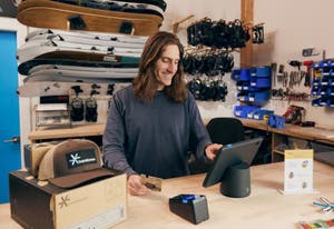 Small business owner checking out a customer purchasing a sweatshirt using the PayPal POS app on an iPad stand with a connected PayPal card reader at a retail counter.