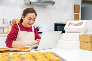 Femme portant un tablier blanc, assise devant un plateau de biscuits, utilisant son ordinateur pour effectuer des ventes en ligne