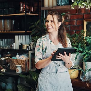 Smiling small business owner in a floral top and linen apron holds a tablet while standing in her plant and homeware shop, surrounded by lush potted greenery, glass vases, and a warm exposed brick wall backdrop